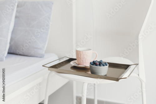 Light breakfast served on a metal tray. Coffee in a pink cup and fresh blueberry on a white chair near bed. Coffee in bed concept © VeronikaSmirnaya