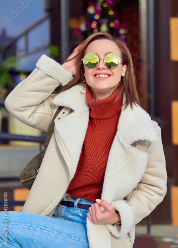 a positive girl in a brown jacket and white sheepskin coat and sunglasses is outdoor.  smile on her face. close up. reflection of the sky in glasses.