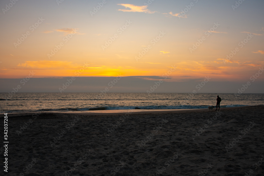 Eine männliche Person läuft mit seinem Hund im orange-roten Sonnenuntergang am Strand entlang Eine männliche Person läuft mit seinem Hund im orange-roten Sonnenuntergang am Strand entlang