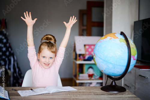 Little caucasian schoolgirl smiling, raised her hands up, reading books, doing homework. Training books, notebooks and globe at the table. Distance learning concept, online education
