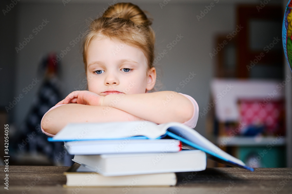 Сute little schoolgirl is tired of reading books at home at the table and is sad. Girl Does Homework. Distance learning Сute little schoolgirl is tired of reading books at home at the table and is sad. Girl Does Homework. Distance learning