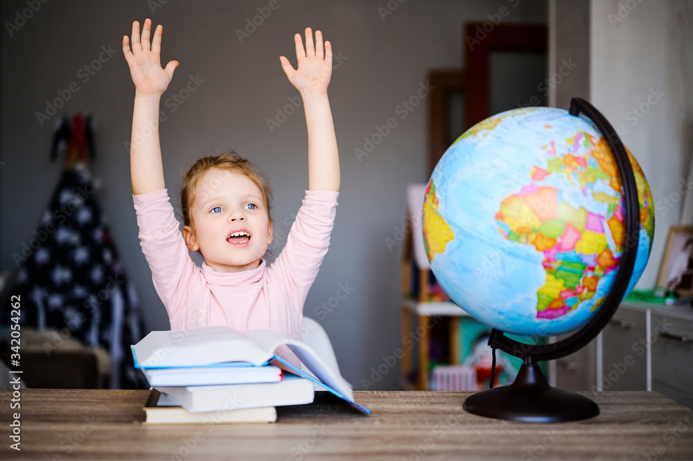 Little caucasian schoolgirl smiling, raised her hands up, reading books, doing homework. Training books, notebooks and globe at the table. Distance learning concept, online education Little caucasian schoolgirl smiling, raised her hands up, reading books, doing homework. Training books, notebooks and globe at the table. Distance learning concept, online education