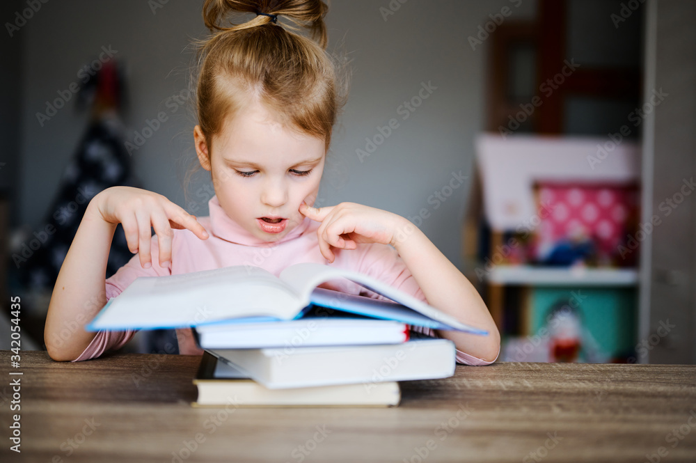 Little girl sitting on the table and reading a book, child doing his homework at home, distance education online learning concept Little girl sitting on the table and reading a book, child doing his homework at home, distance education online learning concept