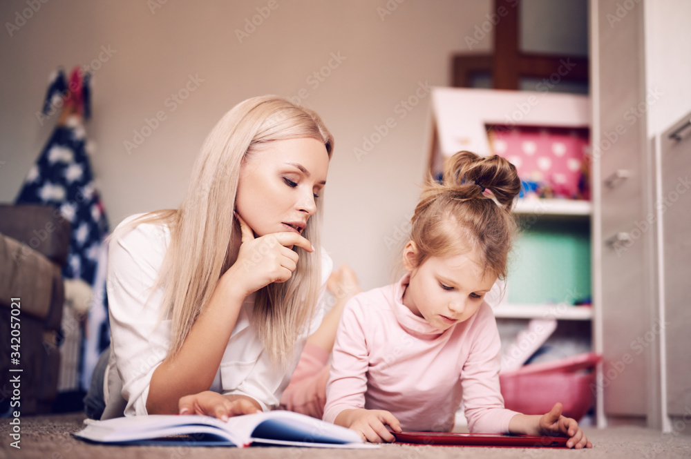 Young mother reads a book at home lying on the floor at home with her daughter, the child watches cartoons on the tablet and plays, happy family, love, family concept. Young mother reads a book at home lying on the floor at home with her daughter, the child watches cartoons on the tablet and plays, happy family, love, family concept.