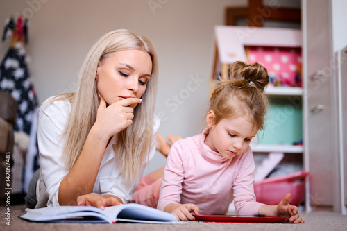 Young mother reads a book at home lying on the floor at home with her daughter  the child watches cartoons on the tablet and plays  happy family  love  family concept.