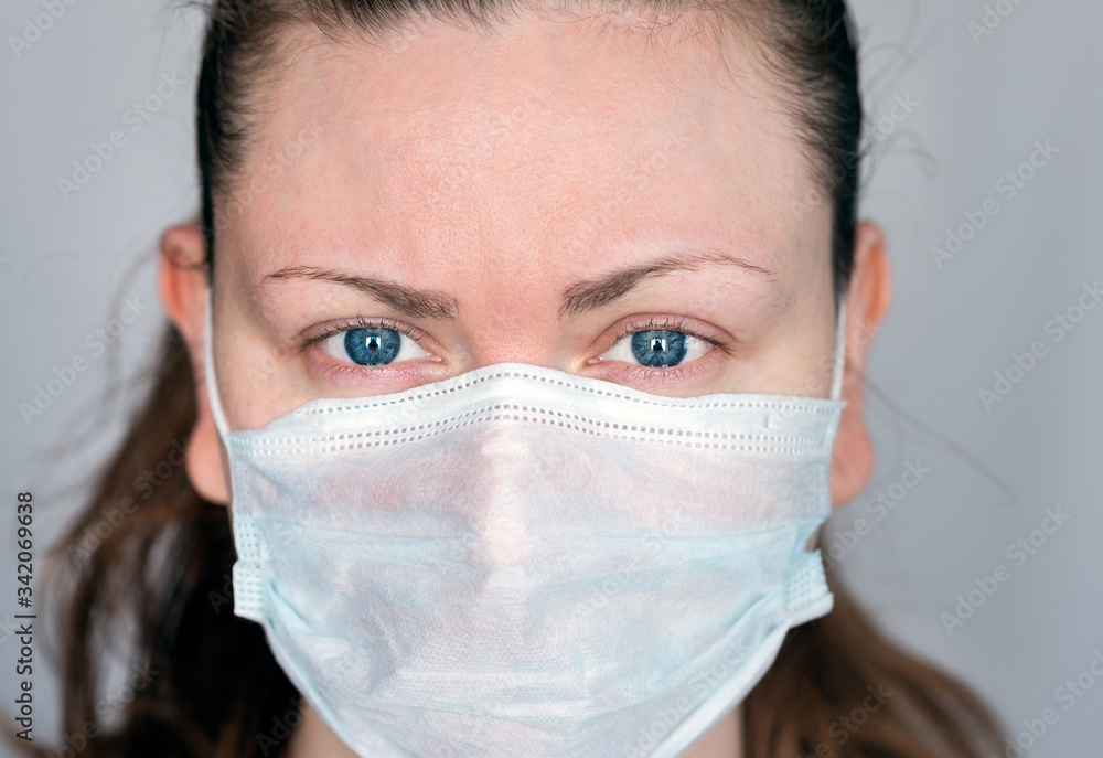 Close up portrait of young woman with blue eyes wearing a surgical mask to protect herself and avoid the spread of covid-19 coronavirus and looking straight at camera - viewer, on gray background Close up portrait of young woman with blue eyes wearing a surgical mask to protect herself and avoid the spread of covid-19 coronavirus and looking straight at camera - viewer, on gray background