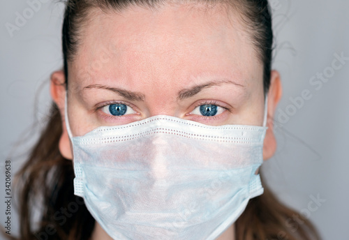 Close up portrait of young woman with blue eyes wearing a surgical mask to protect herself and avoid the spread of covid-19 coronavirus and looking straight at camera - viewer, on gray background photo