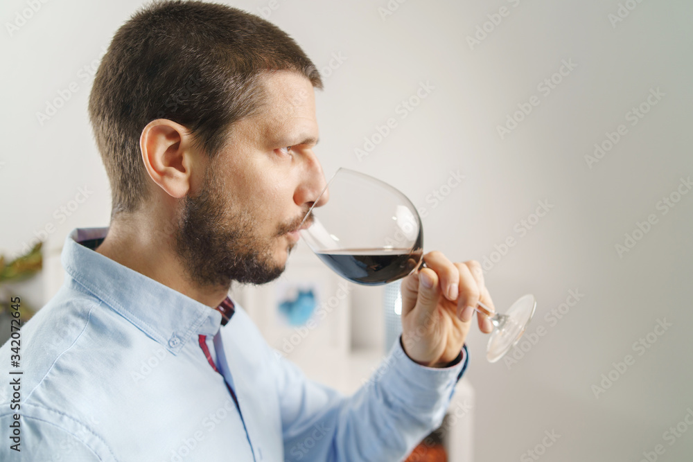 Side view portrait of adult caucasian man wearing shirt holding glass of red wine tasting at home at evening or night close up male with beard and short hair at home alone one Side view portrait of adult caucasian man wearing shirt holding glass of red wine tasting at home at evening or night close up male with beard and short hair at home alone one