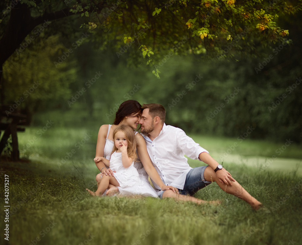 Young barefooted family of three in white summer dresses sitting on a grass in park