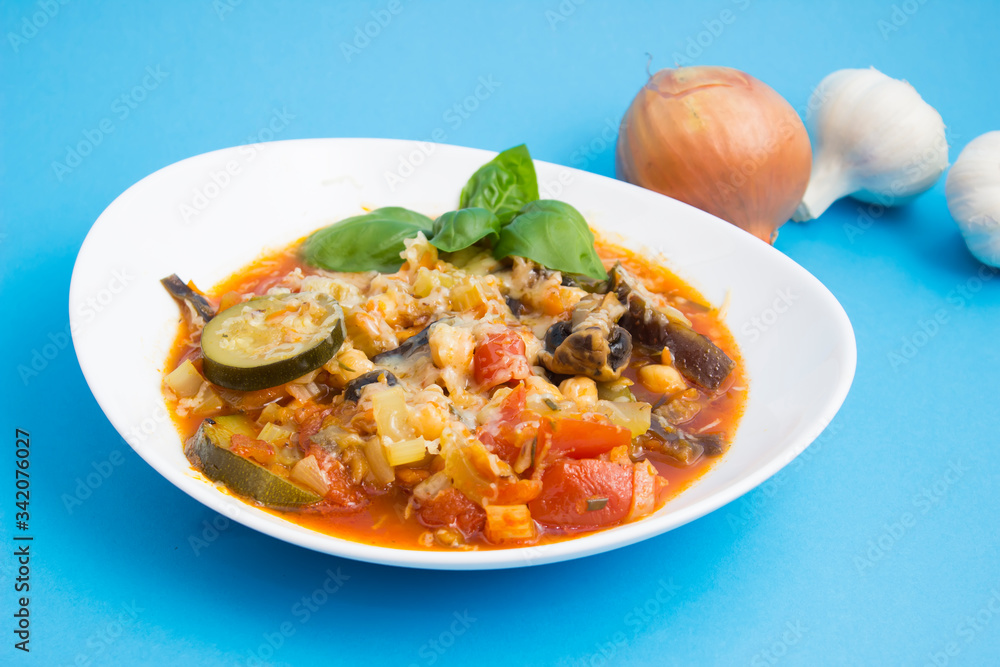 Vegetable stew with eggplant, zucchini, tomatoes and mushrooms. White plate and blue background