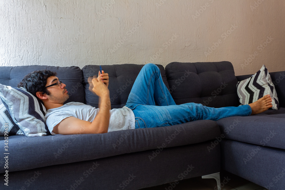 Young Hispanic man with glasses, resting on a sofa, using his cell phone for various things