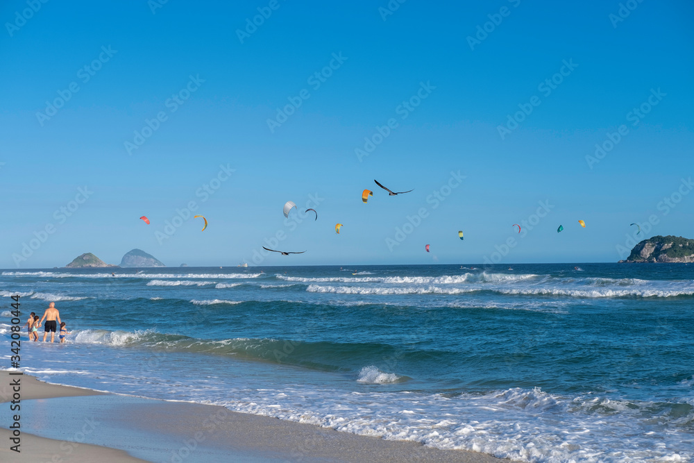Man walks with children on a sandy beach and birds fly over the waves against the background of ocean islands and blue sky on a sunny day. Kite surfing in the ocean on Rio de Janeiro beach in Brasil