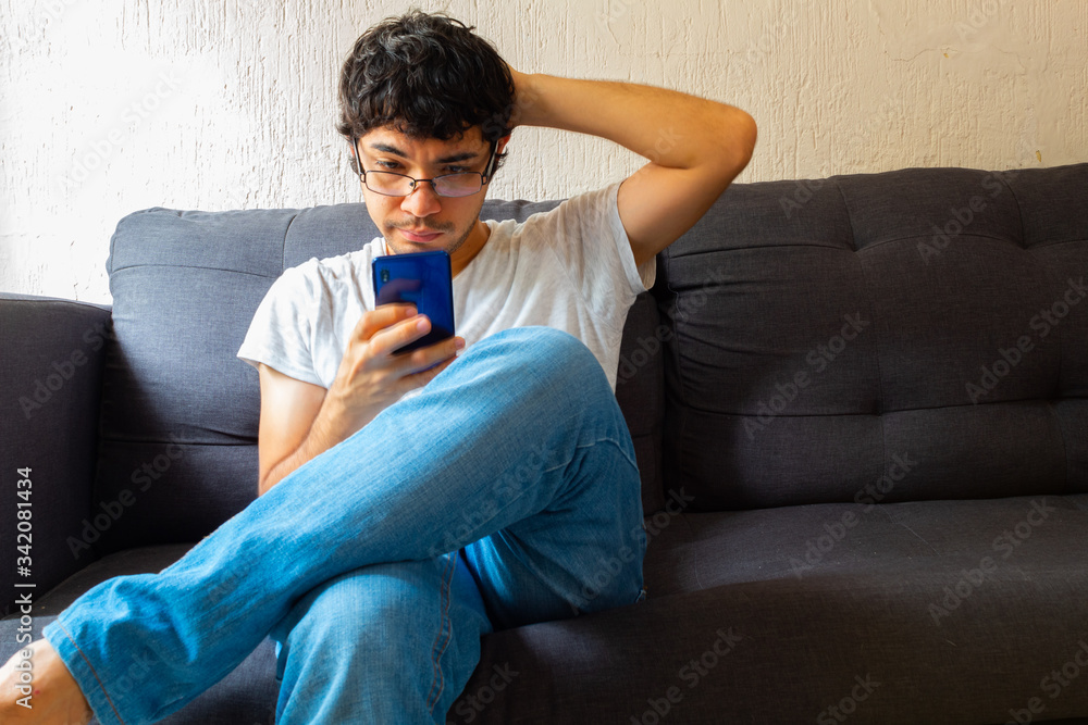 Young Hispanic man with glasses, resting on a sofa, using his cell phone for various things Young Hispanic man with glasses, resting on a sofa, using his cell phone for various things