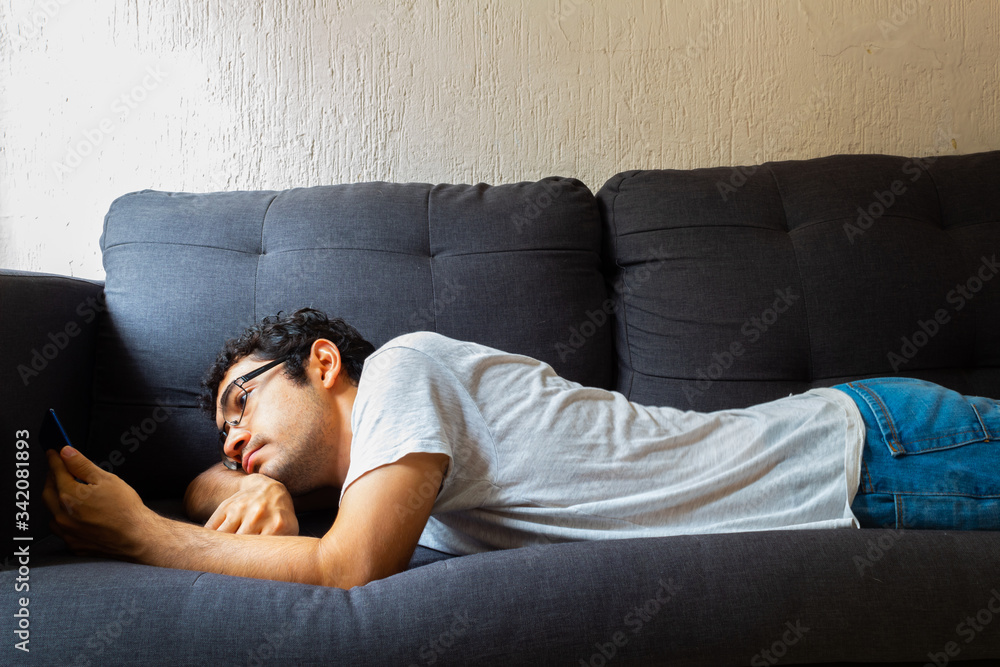 Young Hispanic man with glasses, resting on a sofa, using his cell phone for various things Young Hispanic man with glasses, resting on a sofa, using his cell phone for various things
