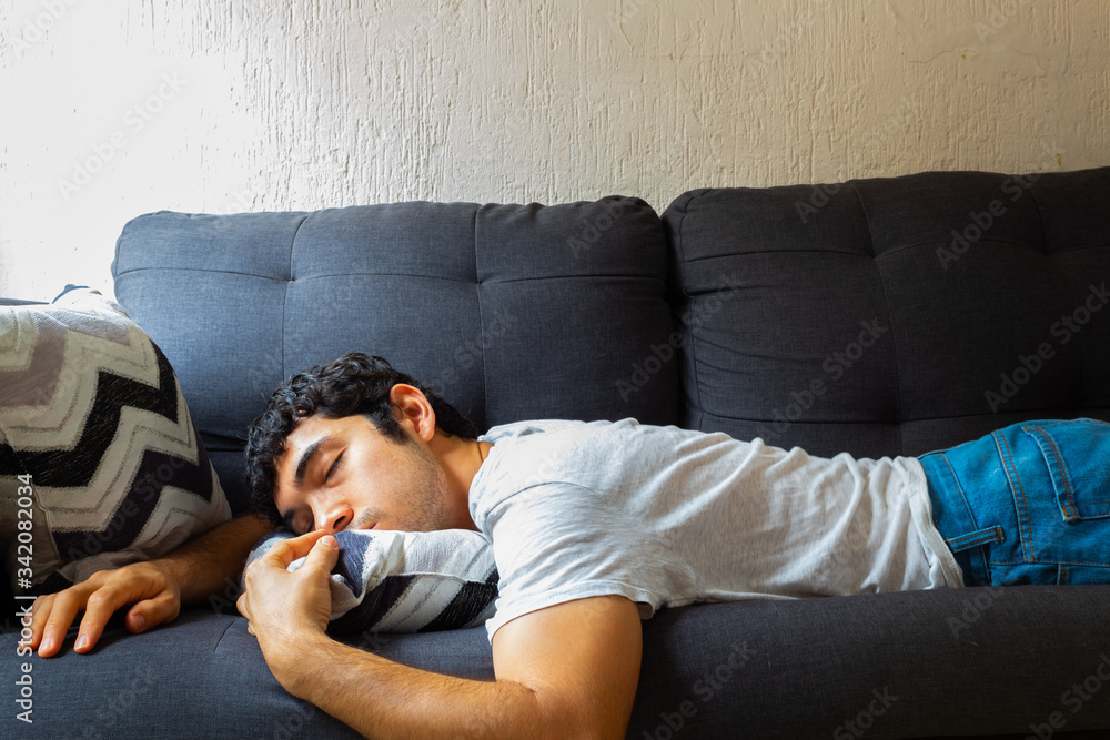 Young Hispanic man with glasses resting on a sofa in the morning, wearing jeans and a white shirt - obrazy, fototapety, plakaty 