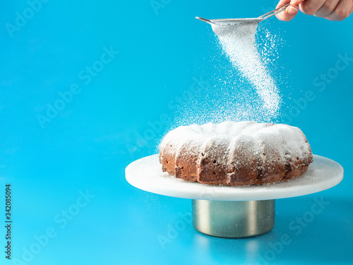 Woman's hand sprinkling icing sugar over fresh home made bundt cake. Powder sugar falls on fresh perfect bunt cake over blue background. Copy space for text. Ideas and recipes for breakfast or dessert