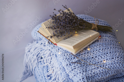 Background of blue hand-knit plaid and pillows in cosy home with dried bouquet of lavender flowers, books, mug of coffee, garland.