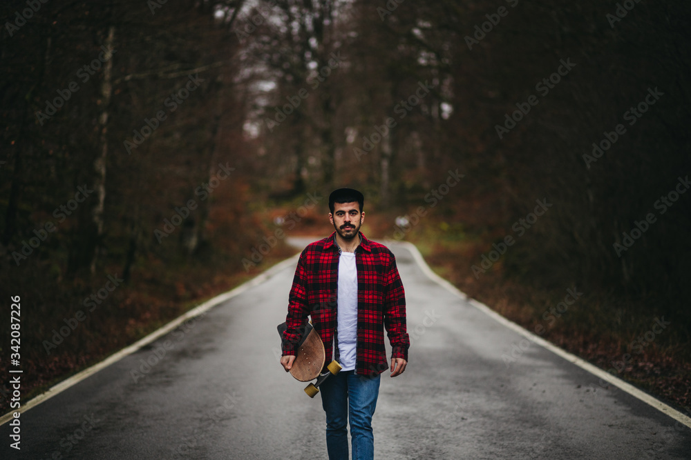 Young stylish man in casual wear walking on asphalt road with skateboard in hand on autumn day looking at camera Young stylish man in casual wear walking on asphalt road with skateboard in hand on autumn day looking at camera