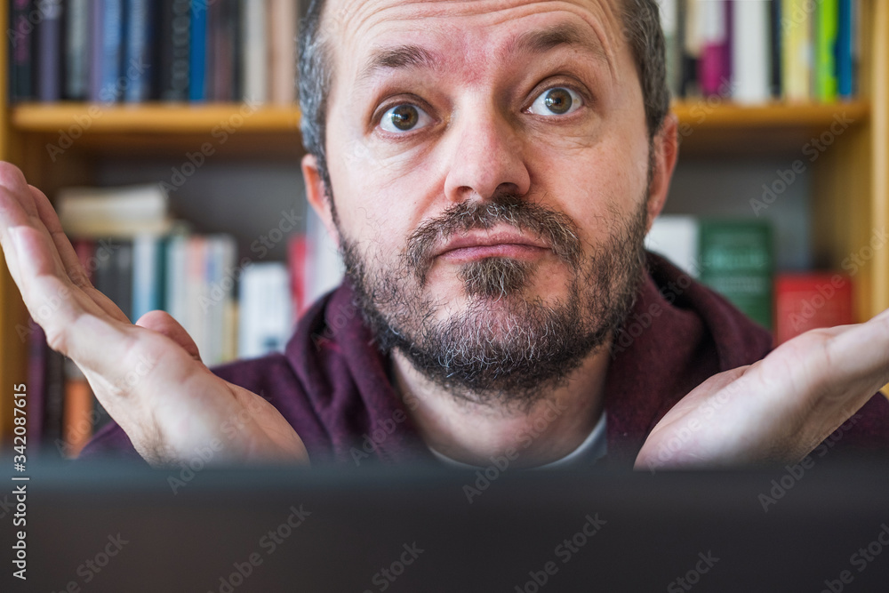 Out of ideas. Confused. Man working from home office. Bearded man working online from home on computer laptop, book shelves behind him. Face expression, confusion. Close up. Out of ideas. Confused. Man working from home office. Bearded man working online from home on computer laptop, book shelves behind him. Face expression, confusion. Close up.