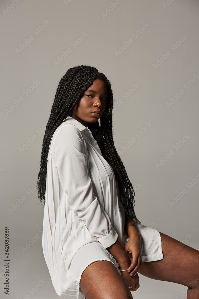 Sensual plus size African American lady with braids in trendy white dress sitting on stool and looking at camera against gray background Sensual plus size African American lady with braids in trendy white dress sitting on stool and looking at camera against gray background