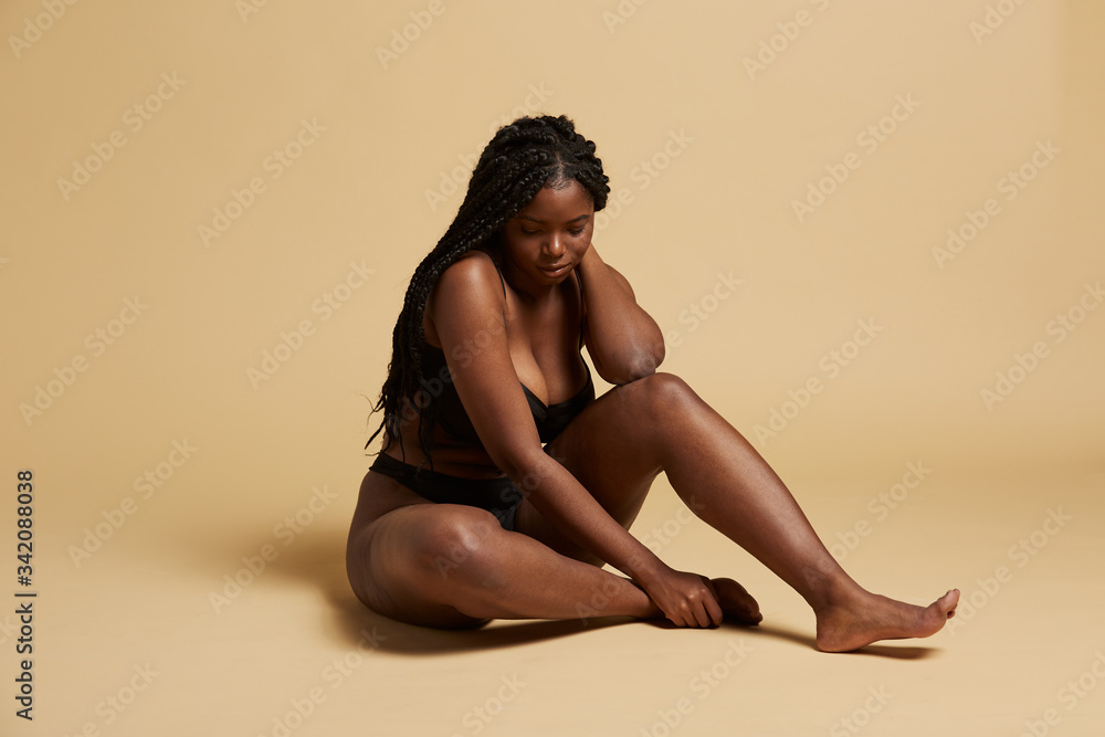 Curvy beautiful African American woman with braids in lingerie looking down sitting on the floor against yellow background Curvy beautiful African American woman with braids in lingerie looking down sitting on the floor against yellow background
