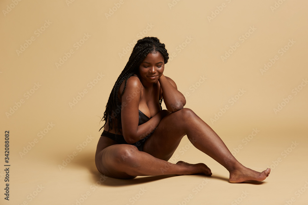 Curvy beautiful African American woman with braids in lingerie looking down sitting on the floor against yellow background Curvy beautiful African American woman with braids in lingerie looking down sitting on the floor against yellow background