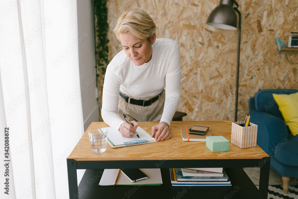 Focused adult businesswoman taking notes of plan on clipboard bending on wooden table in light office Focused adult businesswoman taking notes of plan on clipboard bending on wooden table in light office