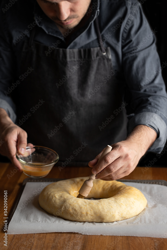 Crop male chef in black apron greasing unbaked round bread with egg yolk while standing at wooden table Crop male chef in black apron greasing unbaked round bread with egg yolk while standing at wooden table