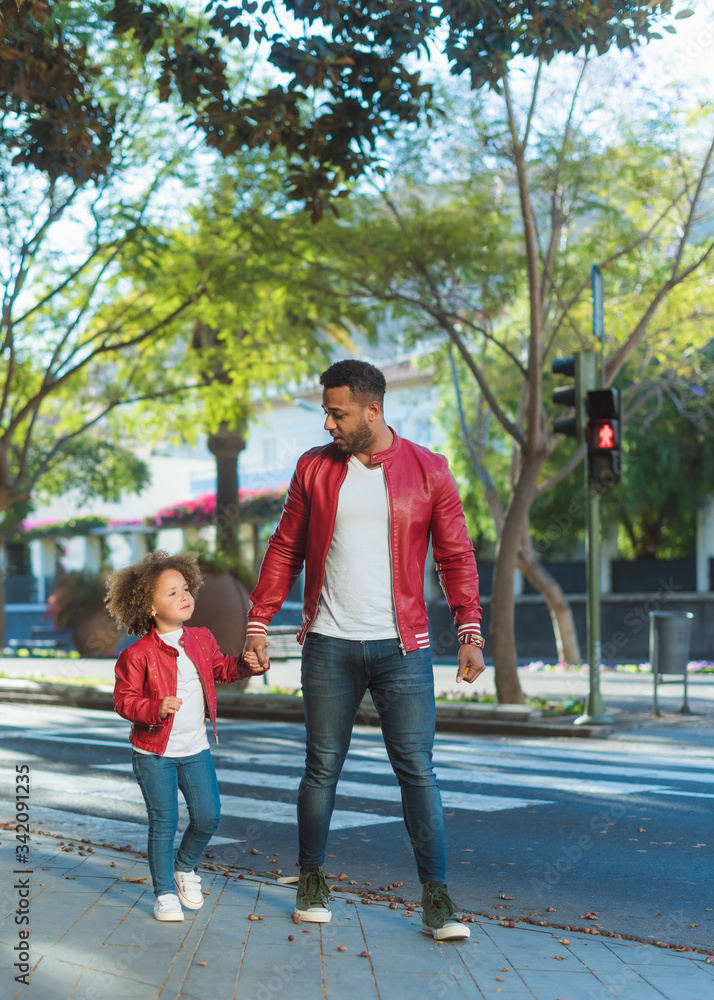 Happy young ethnic man with little daughter dressed in similar outfit holding hands while walking on city street in sunny day Happy young ethnic man with little daughter dressed in similar outfit holding hands while walking on city street in sunny day