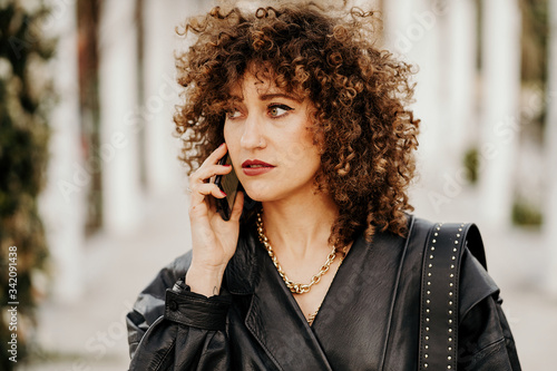 Full body female entrepreneur in vintage outfit walking on asphalt path and having smartphone conversation while commuting to work through city park