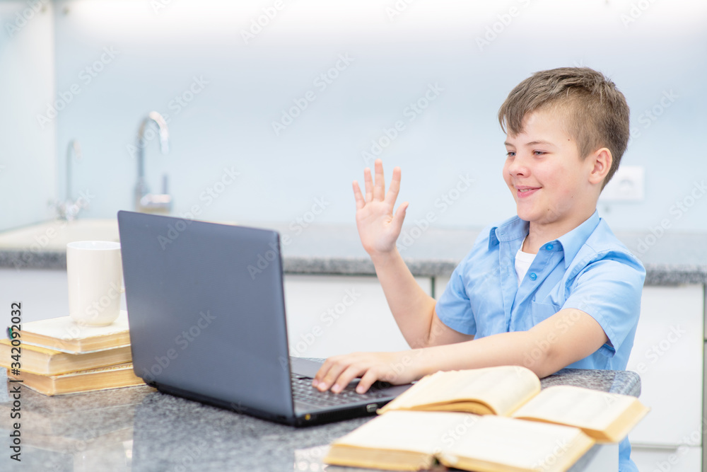 A boy with a blue shirt at home in the kitchen is engaged in education using a laptop. The boy waved his hand in the laptop camera around the child open books laid out. Education during quarantine con A boy with a blue shirt at home in the kitchen is engaged in education using a laptop. The boy waved his hand in the laptop camera around the child open books laid out. Education during quarantine con