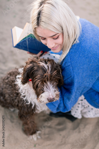 From above happy young female in casual sweater and skirt holding book and stroking adorable curly dog while spending free time on sandy beach photo