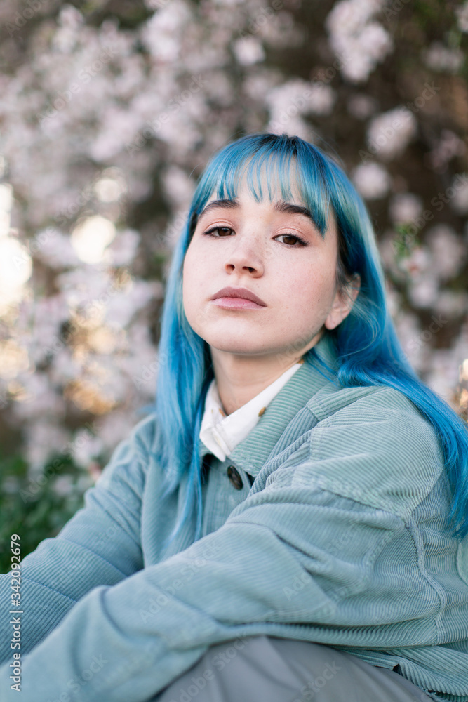 Sad millennial female model with blue hair in stylish outfit looking at camera thoughtfully while sitting on green grass near blooming tree in spring garden