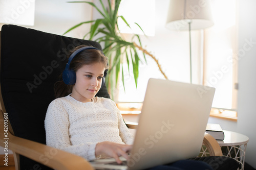 Beautiful 9 year old brunette girl sitting on chair studying on her laptop with headhones photo