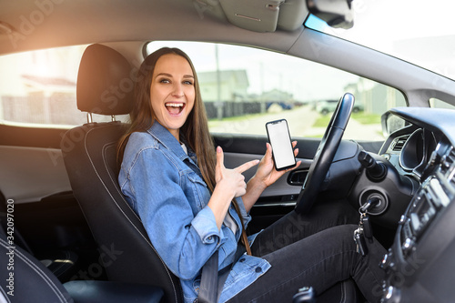 A young beautiful woman driving a car, she points to blank phone screen and smiles. Mobile apps for driver. Inside car view