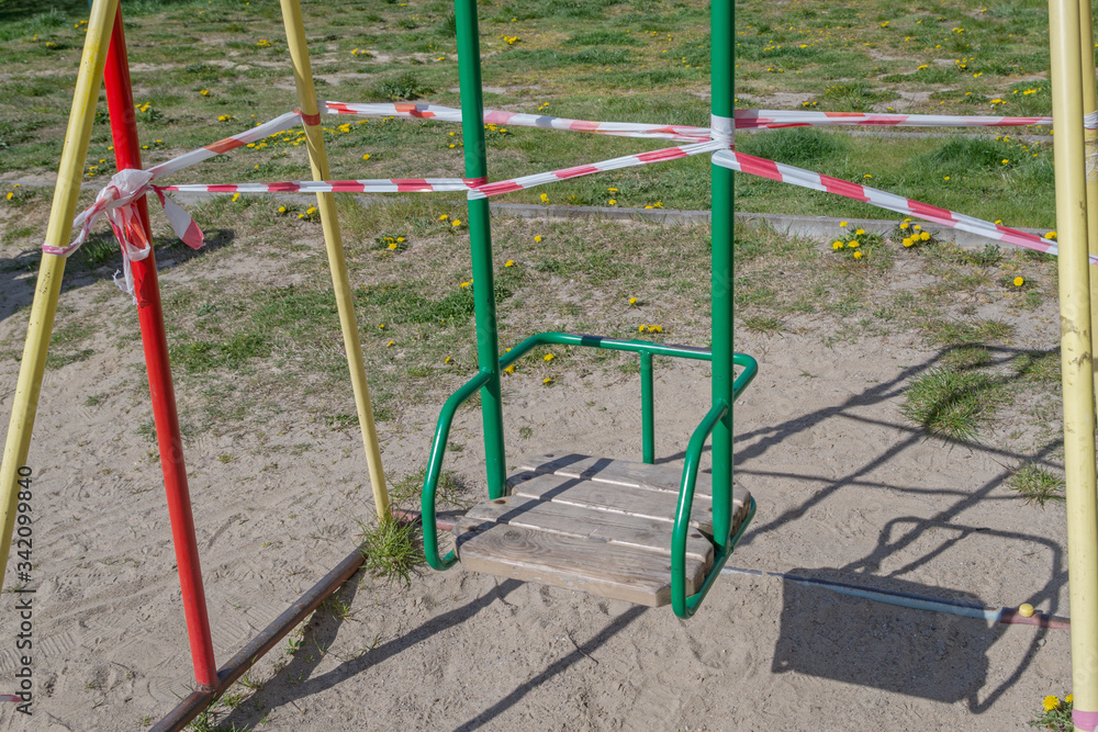 Children's swing with wooden seat with metal green back and handrails, with welded yellow and red iron pipes racks. Swing is tied with red and white signal tape that prohibits use it during quarantine