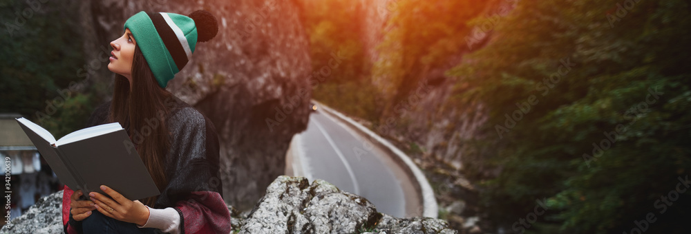 Obraz premium Portrait of young traveler girl reading the book wearing hat of aqua menthe color, on background of asphalting road in mountains.