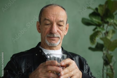 Male portrait of senior man in black leather jacket with glass of brandy indoors in loft style room with light green walls