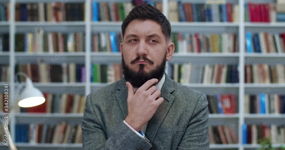 Young Caucasian businessman touching his beard and thinking over something very carefully at books shelves behind. Portrait of serious man considering some philosophy theory in library. Study concept. Young Caucasian businessman touching his beard and thinking over something very carefully at books shelves behind. Portrait of serious man considering some philosophy theory in library. Study concept.