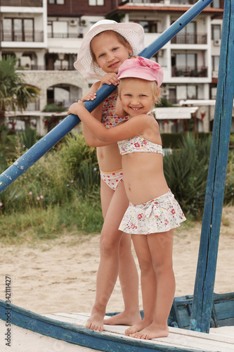 Two little girls in swimsuits and beach hats stand on a boat against the backdrop of hotels and green plants and look into the camera