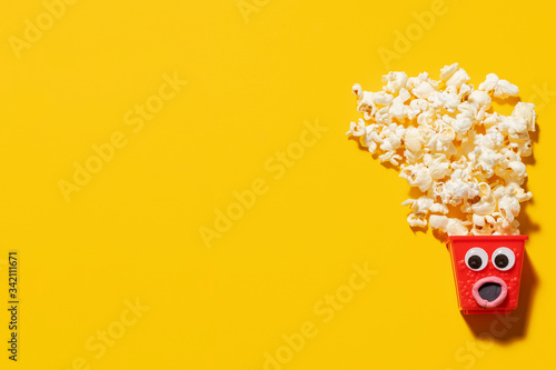Mini red popcorn box carton with googly eyes and a surprised expression with popcorn spilling out the top on a yellow background with copy space and room for text with a right side composition.