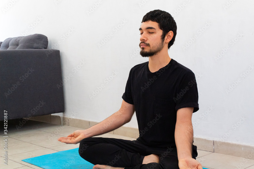 Fototapeta premium young hispanic man wearing black sportswear meditating in a yoga session in the lotus flower pose , at home