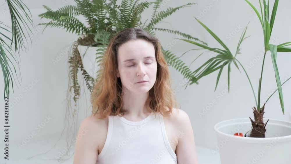 young red headed woman wearing sportswear closes eyes, starts meditating. girl sits in lotus pose, enjoys breath meditation in bright room with house plants at home during quarantine self isolation young red headed woman wearing sportswear closes eyes, starts meditating. girl sits in lotus pose, enjoys breath meditation in bright room with house plants at home during quarantine self isolation