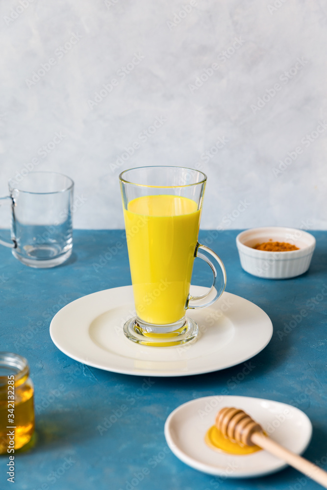 Tall glass with herbal,traditional Indian drink with turmeric stands on white flat plate. Golden milk for disease prevention and immune support.Next to it are ingredients curcum and honey. Copy space Tall glass with herbal,traditional Indian drink with turmeric stands on white flat plate. Golden milk for disease prevention and immune support.Next to it are ingredients curcum and honey. Copy space