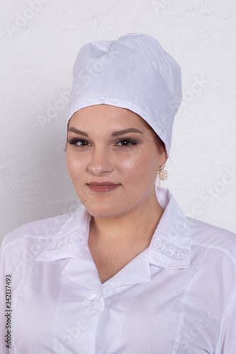 Portrait of attractive young female doctor in white medical jacket and white medical cap on a grey background.