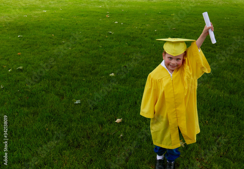 Young boy in a yellow cap and gown smiles and holds up his preschool graduation diploma on a green grass field