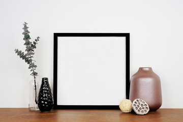 Mock up black square frame with home decor and eucalyptus branch. Wooden shelf against a white wall. Copy space.