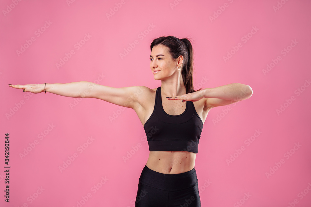 Pumped up sports girl on a pink background in the studio does sports exercises with hands. Smiling and showing her high results by her muscular hands. Fitness for beginners - hands to the side