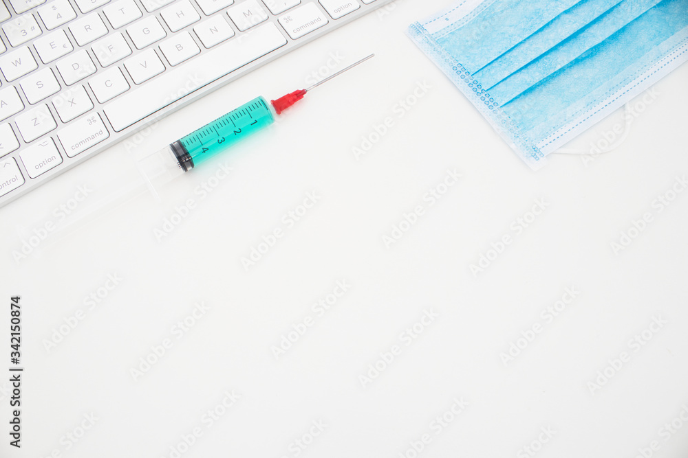 Top view of a composition with a white keyboard, blue face mask and an injection with antiviral drug. White background. Concept of coronavirus, medical research and hope for an antiviral drug Top view of a composition with a white keyboard, blue face mask and an injection with antiviral drug. White background. Concept of coronavirus, medical research and hope for an antiviral drug