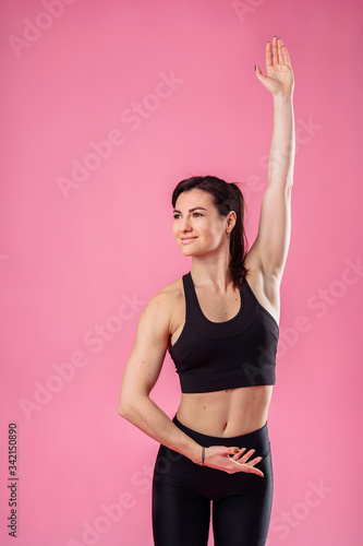 Attractive muscular girl does morning exercises, stretching sweetly, dreaming and smiling. Showing training movements in the studio, isolated on a pink background, wearing black top and pans. © Daria